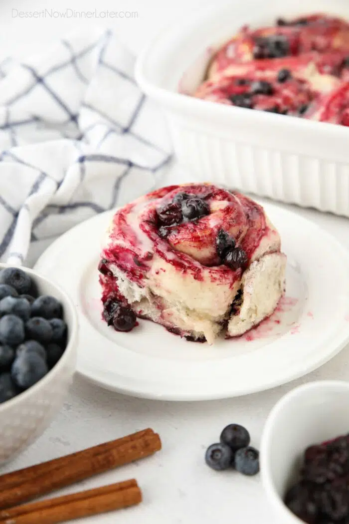 A single blueberry cinnamon roll on a plate with whole blueberries.