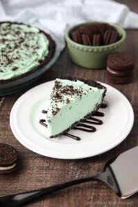 Slice of mint grasshopper pie with an Oreo crust on top of a plate drizzled with chocolate sauce.