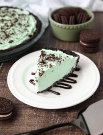 Slice of mint grasshopper pie with an Oreo crust on top of a plate drizzled with chocolate sauce.