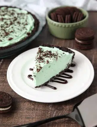 Slice of mint grasshopper pie with an Oreo crust on top of a plate drizzled with chocolate sauce.