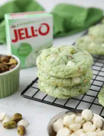 Three pistachio cookies with white chocolate chips stacked on top of each other. Box of pistachio pudding in the background.