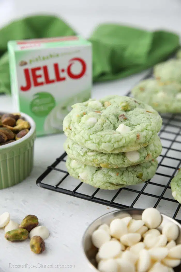 Three pistachio cookies with white chocolate chips stacked on top of each other. Box of pistachio pudding in the background.