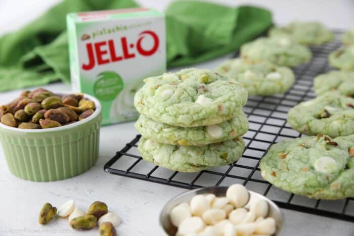 Stack of pistachio pudding cookies on a cooling rack with a jello pudding box, pistachio nuts, and white chocolate chips nearby.