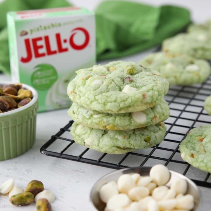 Stack of pistachio pudding cookies on a cooling rack with a jello pudding box, pistachio nuts, and white chocolate chips nearby.