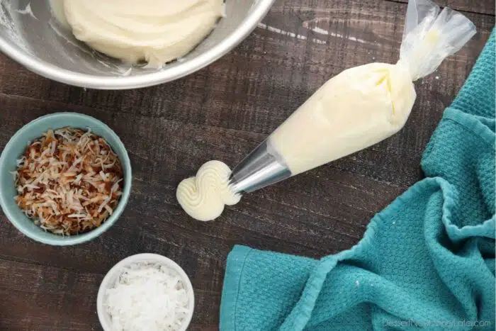 Piping bag with frosting coming out and two bowls of toasted coconut and sweetened coconut flakes.