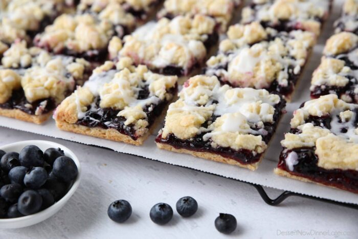 Close up of dessert bars with blueberry pie filling sandwiched between a crust and crumb topping with glaze drizzled on top.