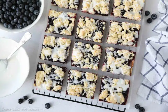 Dessert bars with blueberry pie filling sandwiched between a crust and crumb topping with glaze drizzled on top.
