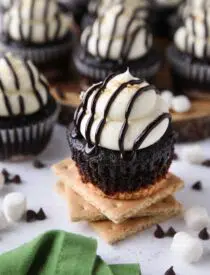 S'mores Cupcakes made with a graham cracker crust, chocolate cake, and marshmallow buttercream frosting topped with chocolate sauce and crushed graham crackers.