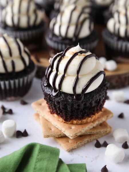 S'mores Cupcakes made with a graham cracker crust, chocolate cake, and marshmallow buttercream frosting topped with chocolate sauce and crushed graham crackers.
