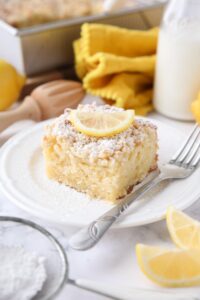 Thick slice of lemon coffee cake on a plate dusted with powdered sugar and a slice of lemon on top.