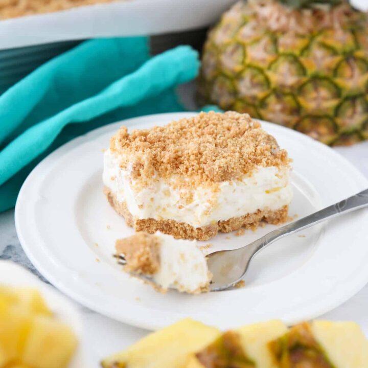 A slice of pineapple delight on a plate with a fork-full taken out. A creamy pineapple dream dessert with graham crackers, cream cheese, and whipped topping.