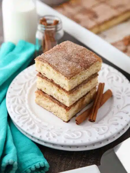 Stack of sopapilla cheesecake bars on a plate with cinnamon sticks.