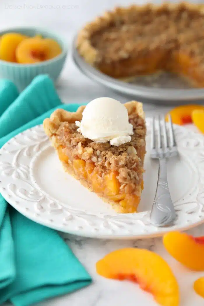 Slice of peach crumb pie on a plate with vanilla ice cream on top.