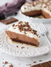 Slice of chocolate truffle pie on a plate showing a graham cracker crust and layers of ganache, whipped chocolate, whipped cream, and chocolate shavings on top.