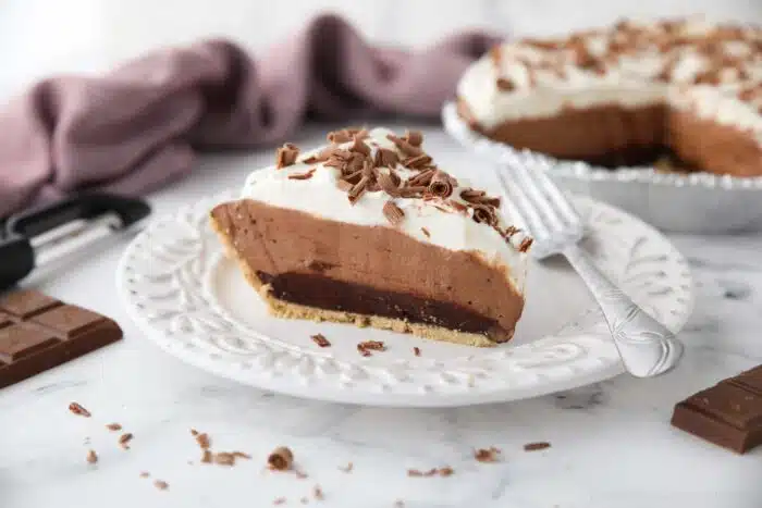 Slice of chocolate truffle pie on a plate showing a graham cracker crust and layers of ganache, whipped chocolate, whipped cream, and chocolate shavings on top.