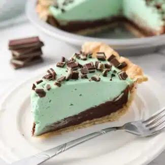 Slice of chocolate mint pie on a plate with layers of pastry crust, chocolate ganache, mint no bake cheesecake, and chopped Andes mints.