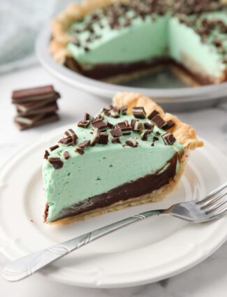 Slice of chocolate mint pie on a plate with layers of pastry crust, chocolate ganache, mint no bake cheesecake, and chopped Andes mints.