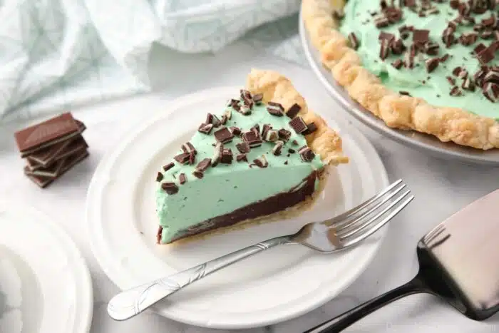 Slice of chocolate mint pie on a plate with layers of pastry crust, chocolate ganache, mint no bake cheesecake, and chopped Andes mints.