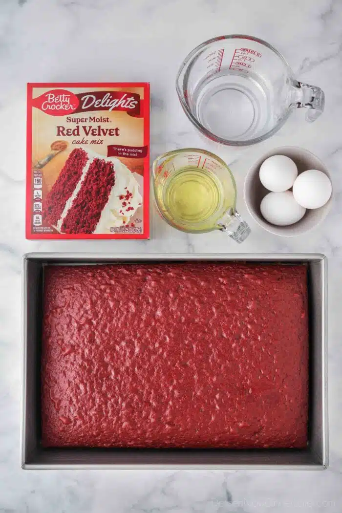 Box of Red Velvet Cake Mix and ingredients needed to make it with the finished cake in a 13x9-inch pan.
