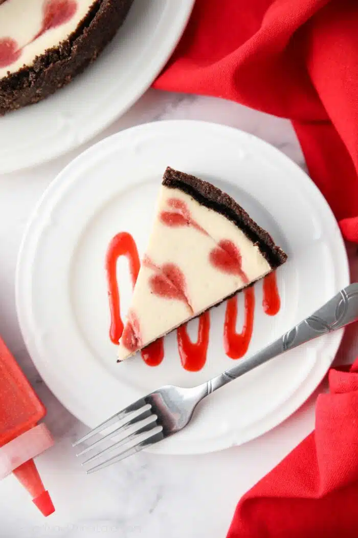 Strawberry sauce drizzled on a plate with a slice of Valentine's Day Cheesecake on top with red hearts baked into the batter.