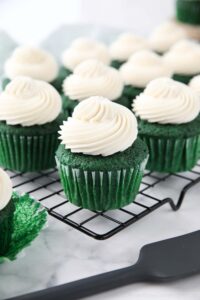 Cooling rack full of green cupcakes with whipped cream cheese frosting piped on top.