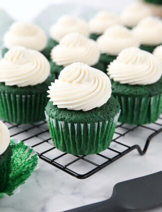 Cooling rack full of green cupcakes with whipped cream cheese frosting piped on top.