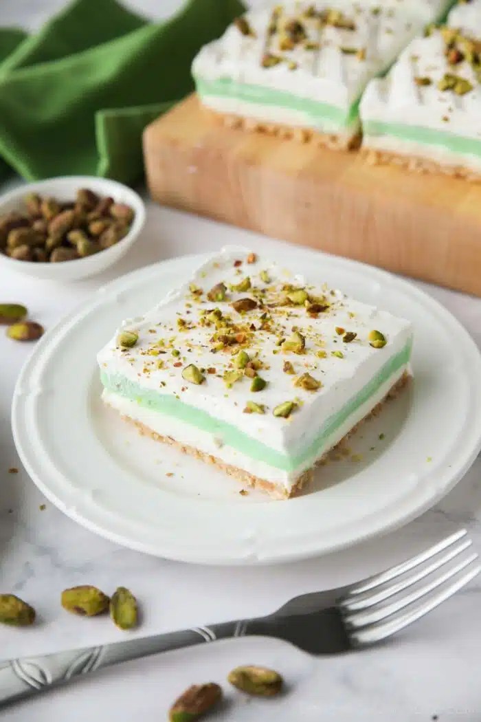 Layered dessert with a shortbread cookie crust, no-bake cheesecake layer, pistachio pudding layer, and whipped cream on top sprinkled with chopped pistachios.