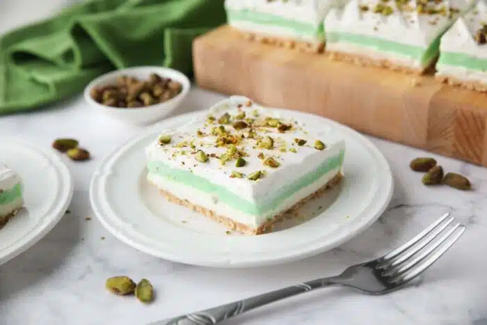 Layered dessert with a shortbread cookie crust, no-bake cheesecake layer, pistachio pudding layer, and whipped cream on top sprinkled with chopped pistachios.