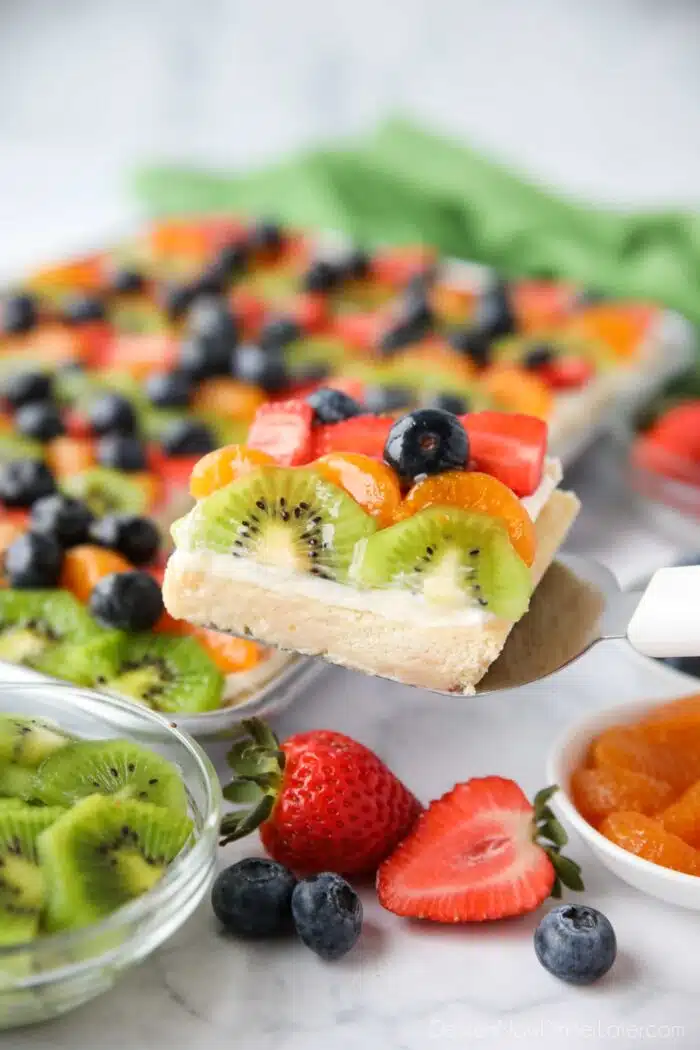 Scooping a sugar cookie fruit pizza bar with cream cheese frosting from tray.