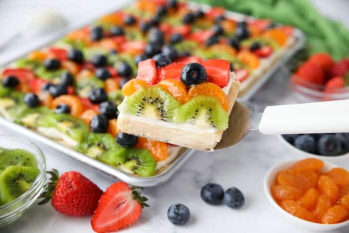 Scooping a sugar cookie fruit pizza bar with cream cheese frosting from tray.