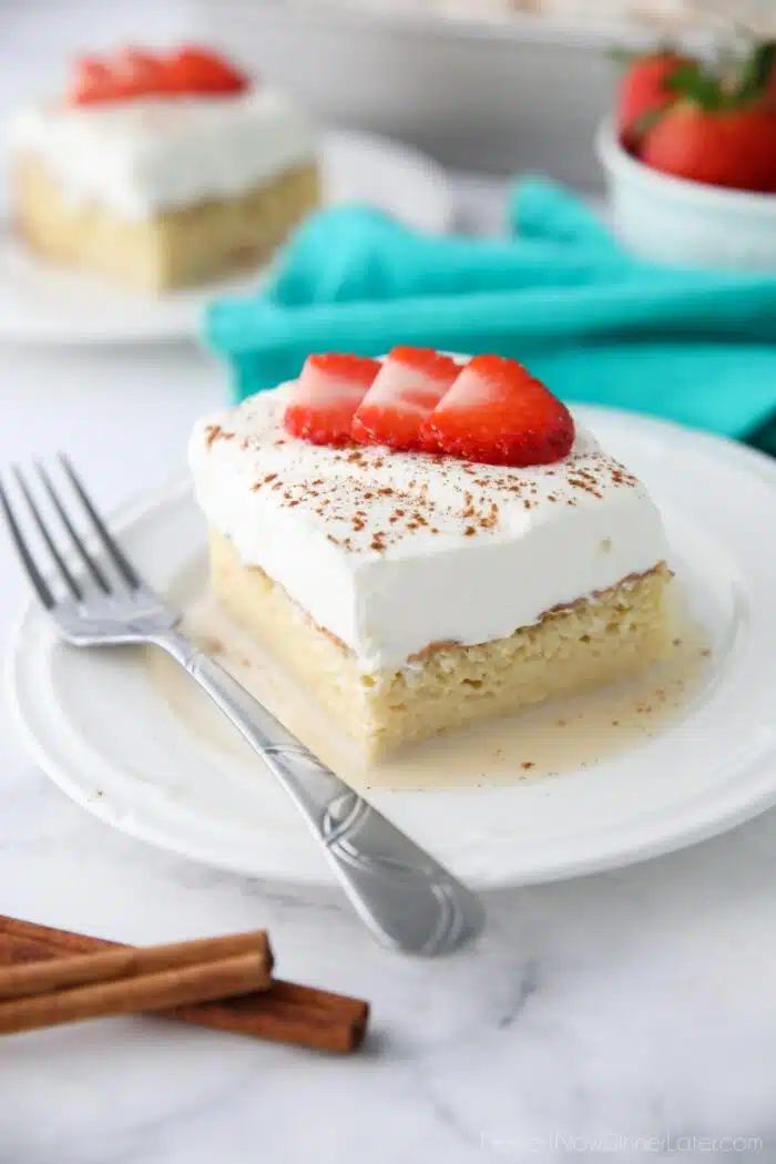 Slice of tres leches cake on a plate with whipped cream, cinnamon, and sliced strawberries on top.