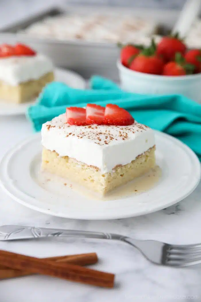 Slice of tres leches cake on a plate with whipped cream, cinnamon, and sliced strawberries on top.