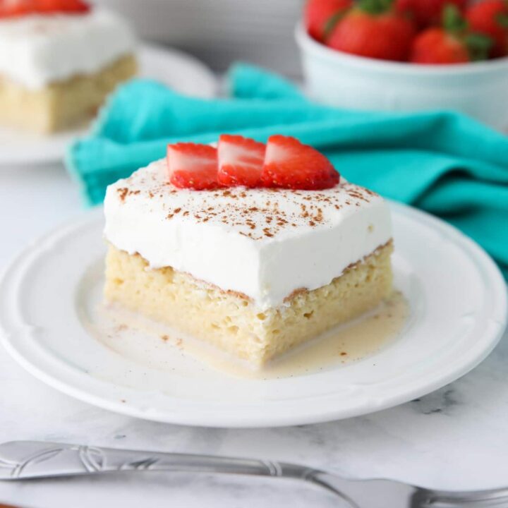 Slice of tres leches cake on a plate with whipped cream, cinnamon, and sliced strawberries on top.