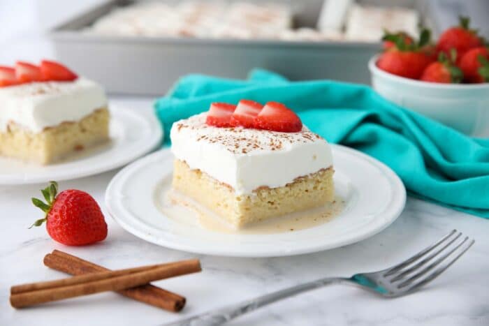 Slice of tres leches cake on a plate with whipped cream, cinnamon, and sliced strawberries on top.