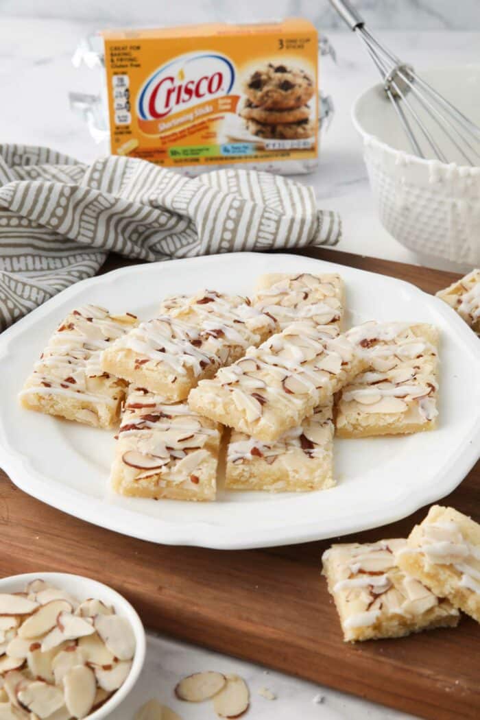 Side view of almond bars on a plate with Crisco shortening, sliced almonds, and a bowl of glaze nearby.