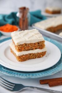 Two carrot cake bars with frosting stacked on a plate.