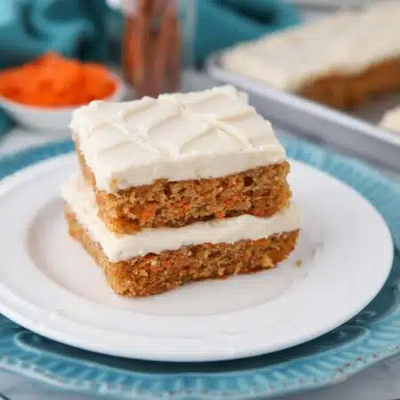Two carrot cake bars with frosting stacked on a plate.