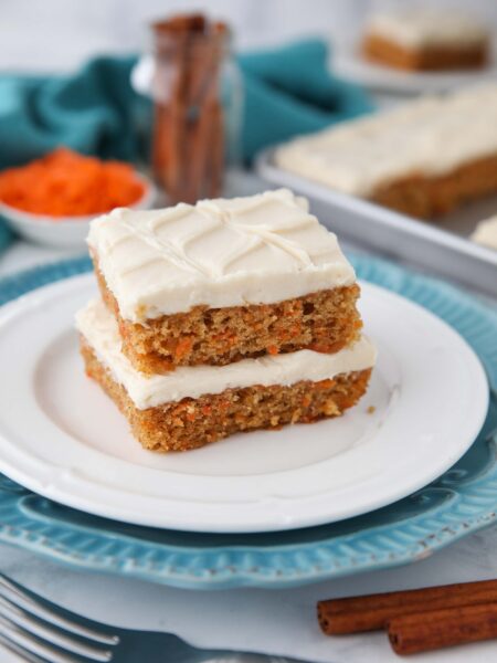 Two carrot cake bars with frosting stacked on a plate.