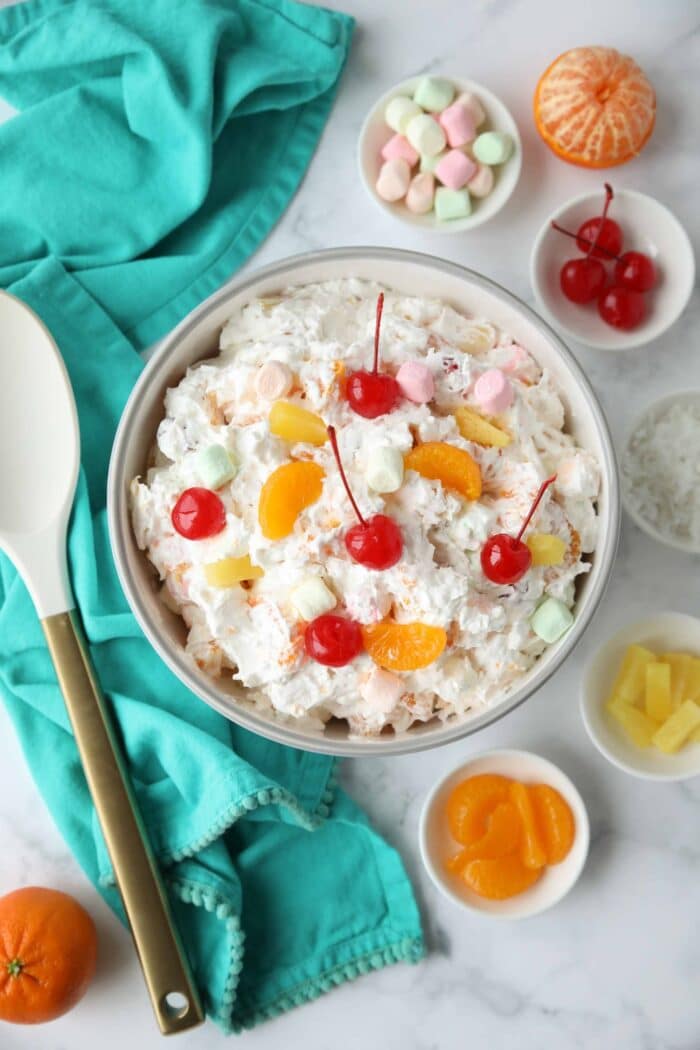 Top view of ambrosia salad in a bowl with canned mandarin oranges, pineapple tidbits, maraschino cherries, shredded coconut, mini marshmallows, Greek yogurt and Cool Whip.