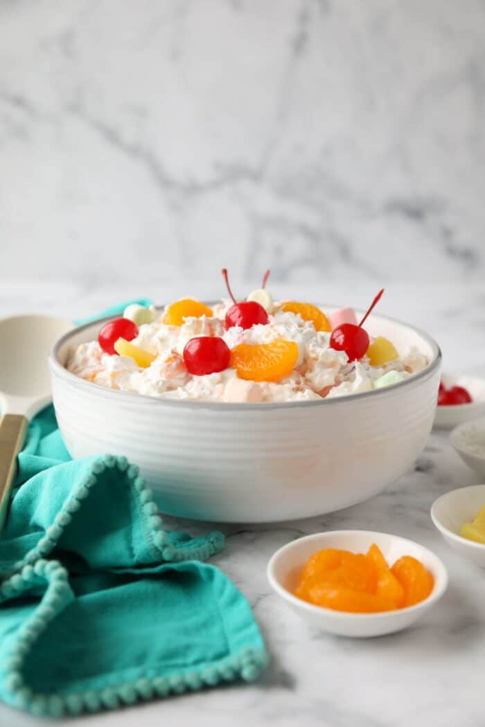 Side view of ambrosia salad in a bowl with canned mandarin oranges, pineapple tidbits, maraschino cherries, shredded coconut, mini marshmallows, Greek yogurt and Cool Whip.