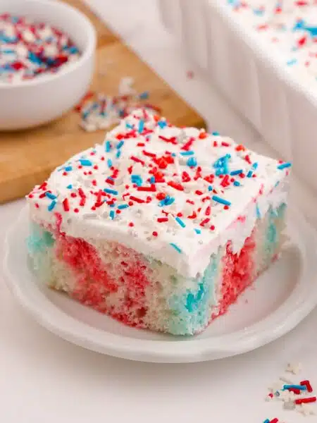 Red white and blue poke cake on a plate with Cool Whip and patriotic sprinkles on top.