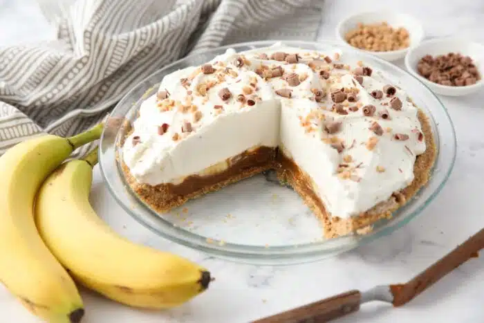 Slices removed from banoffee pie showing layers of graham cracker crust, dulce de leche caramel, bananas, whipped cream and toppings.