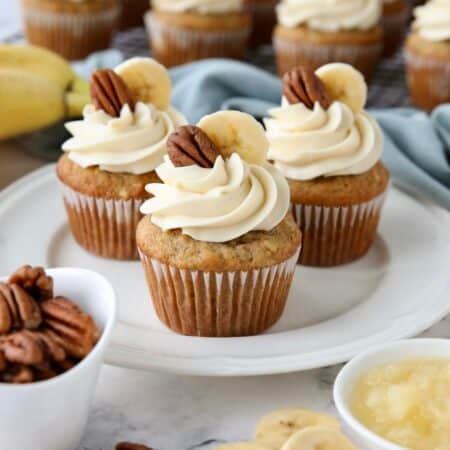 Three hummingbird cupcakes on a plate with cream cheese frosting and a slice of banana and pecan on top.